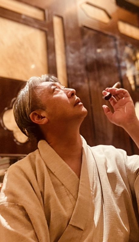 ローブ姿で葉巻を手に天を仰ぐ西田理一郎