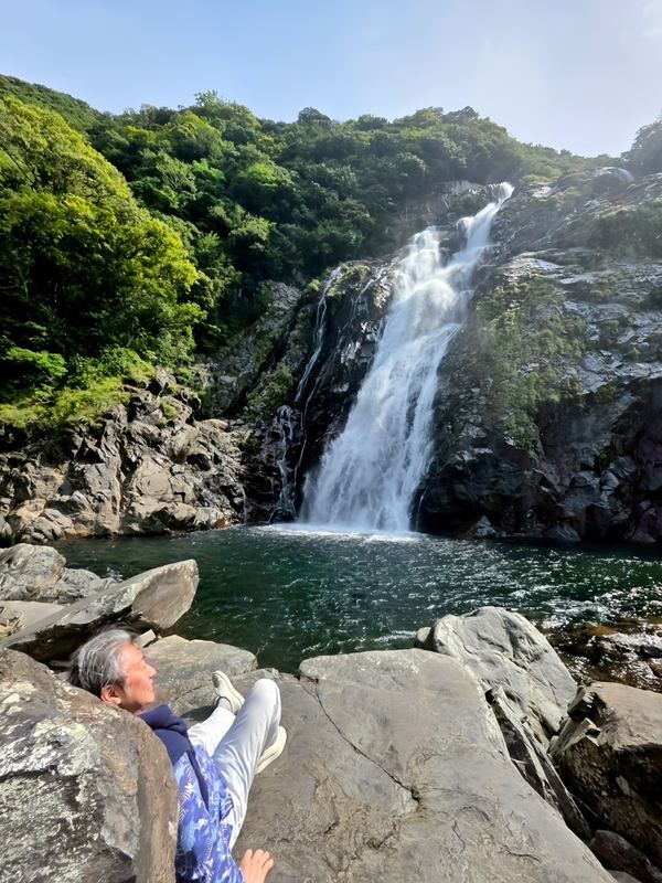 滝のそばの岩に腰かける西田理一郎