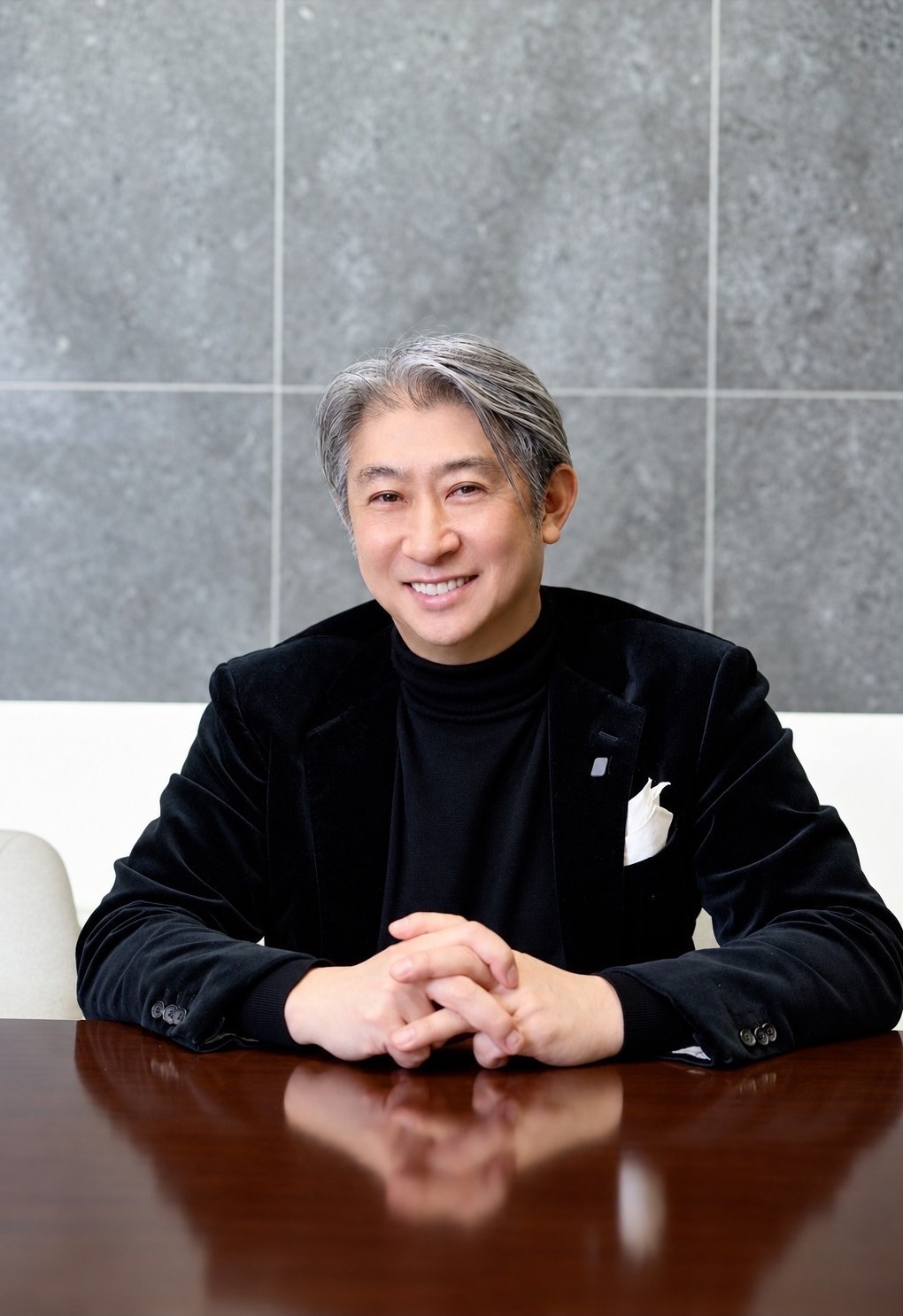 黒のベルベットのジャケットで、卓に手を組んで笑う西田理一郎